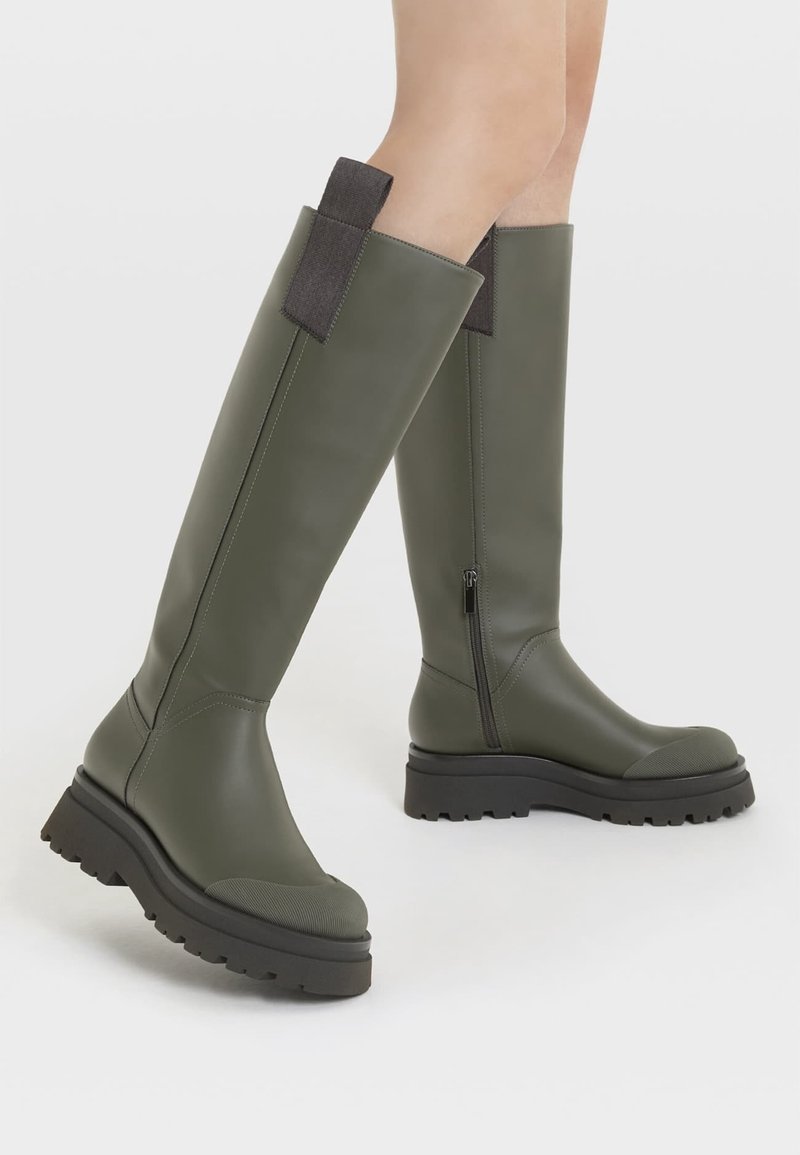 Bottes vert olive montant jusqu'au genou avec semelles noires épaisses, fermeture éclair sur le côté intérieur et languettes en tissu, portées sur jambes nues.