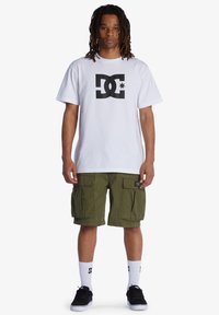 Weißes Baumwoll-T-Shirt mit schwarzem DC-Logo, olivgrüne Cargo-Shorts mit Taschen, weiße Socken mit Logo und schwarze Skate-Schuhe.