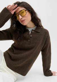 Pull en laine marron avec un col boutonné, des poignets côtelés et une finition texturée, associé à de grandes lunettes de soleil jaunes.