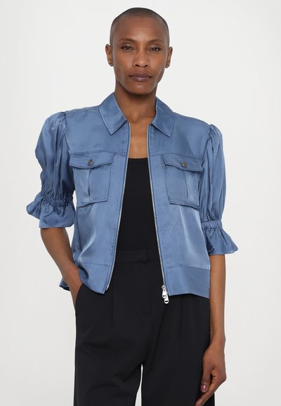 Gaiši zilas satīna jaka ar rāvējslēdzēju un īsām puffed piedurknēm, divām priekšējām kabatām un apkakles izgriezumu. Gluda tekstūra ar smalku spīdumu.