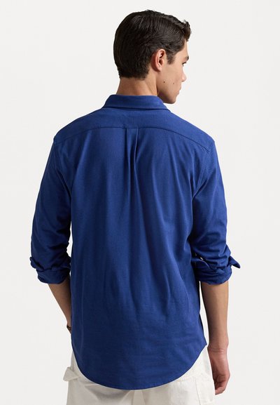 Blauw langemouwen shirt gemaakt van zachte stof met een puntige kraag, opgerolde mouwen en een afgeronde zoom. Bevat een rugpas en plooien.
