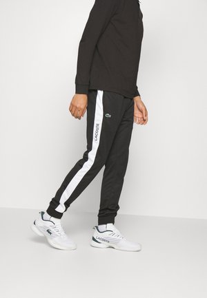 Person iført sorte Lacoste træningsbukser med hvide striber og hvide Lacoste sneakers, gående mod en ensfarvet lys baggrund.