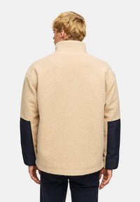 Beige fleece-jacka med hög krage och marinblåa tygdetaljer på ärmarna. Mjuk textur; avslappnad passform.
