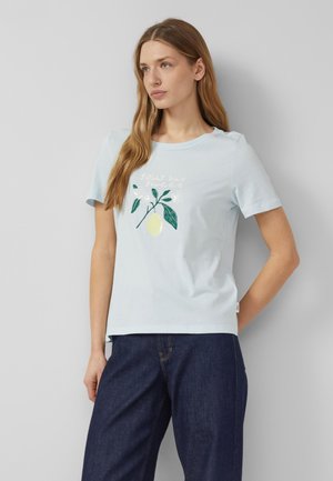 Młoda kobieta z długimi włosami, ubrana w jasnoniebieską koszulkę z nadrukiem cytryny i napisem „kwaśne, ale słodkie”, w ciemnoniebieskich dżinsach, stojąca przy ścianie.