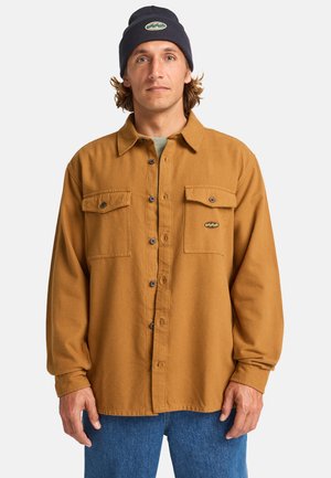 Brown flannel shirt with two chest pockets, button closure, and logo patch. Worn with a dark blue beanie and blue denim pants.