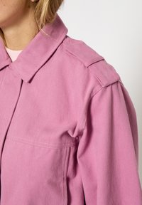 Rozā kokvilnas jaka ar apkakli, plecu uzplečiem un sānu kabatas detaļām. Audums šķiet struktūrēts un izturīgs.