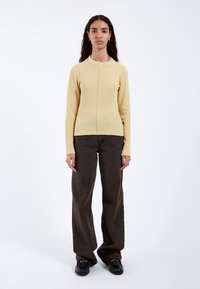 Light yellow, ribbed long-sleeve top with a round neckline, paired with dark brown wide-leg jeans and black shoes, standing against a white background.