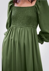 Abito verde con corpo arricciato, scollatura quadrata e lunghe maniche a sbuffo. Il tessuto è morbido, con una texture liscia e un design fluido.