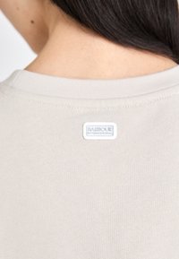 Beige színű pamut póló, kerek nyakkivágással, a hátán egy kis fehér téglalap alakú címkével, amelyen a "BARBOUR" felirat található. Sima textúra.