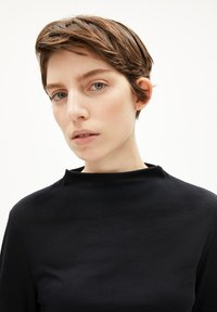 Schwarzes Oberteil mit hohem Kragen und glatter Textur; der Stoff wirkt leicht. Das Model hat kurzes braunes Haar und hellblaue Augen.