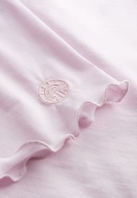 Light pink cotton fabric with a ruffled edge. Features a small, embroidered circular logo in a matching light pink thread.