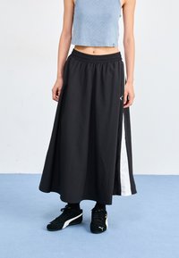 Svart maxi skjørt med elastisk liv, med en hvit sidestripe. Brukt med svarte sneakers. Bakgrunnen er lyseblå.
