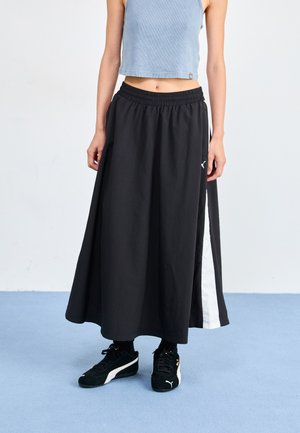Jupe maxi noire avec une taille élastique, dotée d'une bande latérale blanche. Portée avec des baskets noires. Fond de couleur bleu clair.