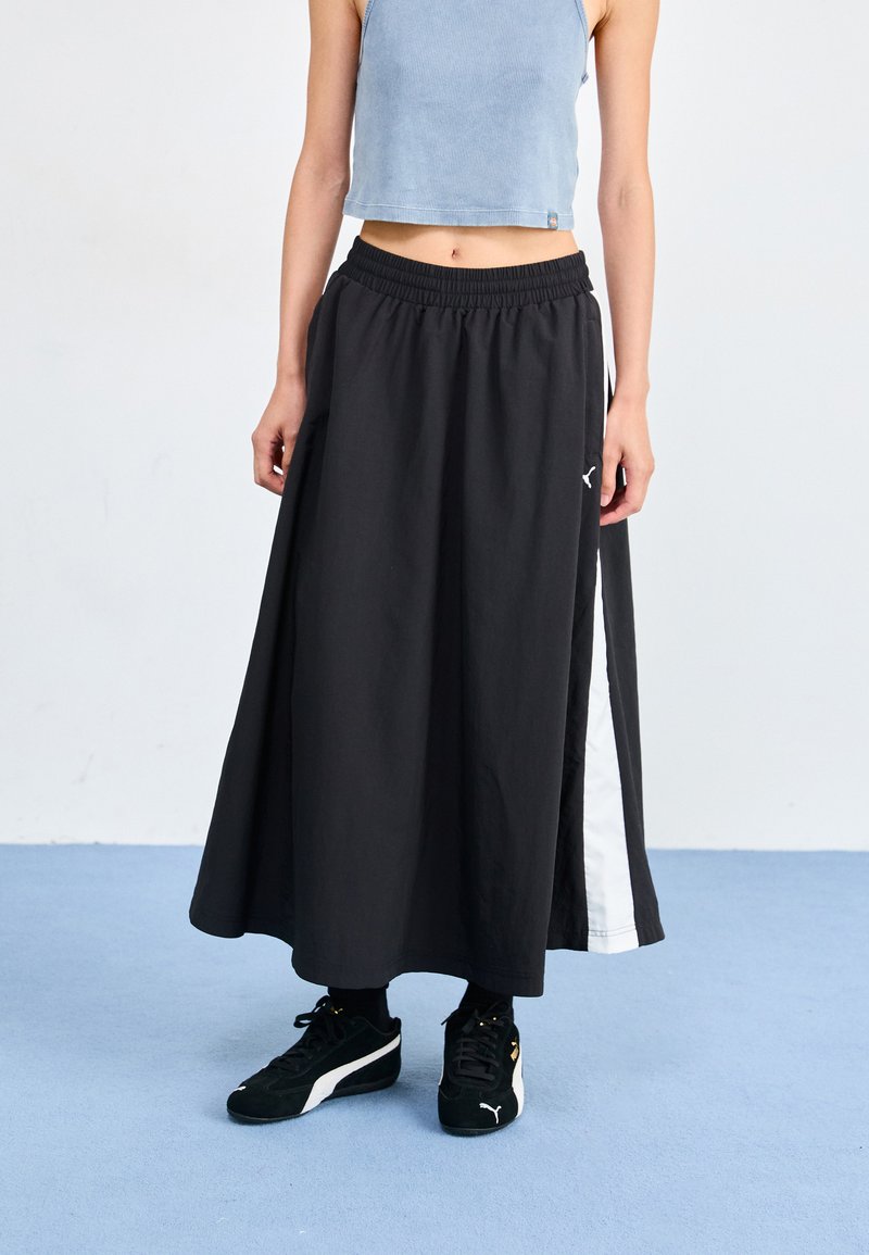 Svart maxi skjørt med elastisk liv, med en hvit sidestripe. Brukt med svarte sneakers. Bakgrunnen er lyseblå.