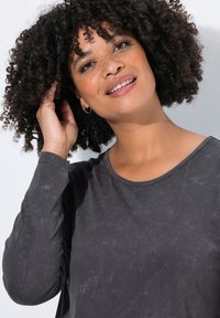 Dunkelgraues Langarm-T-Shirt mit einem runden Ausschnitt und strukturiertem Stoff. Modell mit lockigem Haar posiert und berührt leicht ihr Haar.
