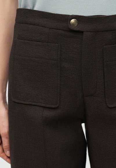 Κοντινή λήψη μαύρου υφασμάτινου παντελονιού με εμπρόσθιες τσέπες με επιδιόρθωση και κλείσιμο με ορείχαλκο κουμπί, φορεμένο με γαλάζιο τοπ.