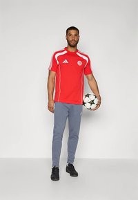 Sarkana sporta krekls ar īsām piedurknēm un baltām detaļām un logo, kombinēts ar pelēkām sporta biksēm. Modelis tur rokās futbola bumbu un valkā melnas kurpes.