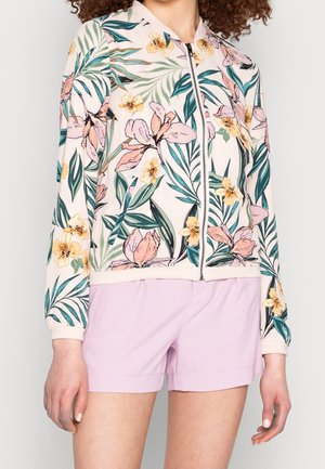 Femme portant une veste zippée avec des motifs floraux verts et roses et un short violet clair, montrée du cou jusqu'à la mi-cuisse.