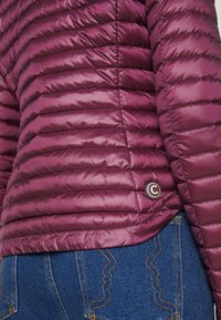 Bordeaux pufferjacka med horisontell quiltad textur och en rund logotypdetalj. Bärs över mörkblå jeans med kontrasterande sömmönster.