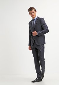 Costum tailor-made de culoare bleumarin, realizat dintr-un material neted, având un sacou cu două butoane, pantaloni slim-fit și o cămașă bleu deschis cu cravată.