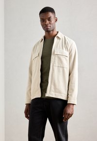 Roomwitte corduroy jas met twee borstzakken, gedragen over een donkergroen t-shirt, gecombineerd met zwarte jeans. Eenvoudig, functioneel ontwerp.
