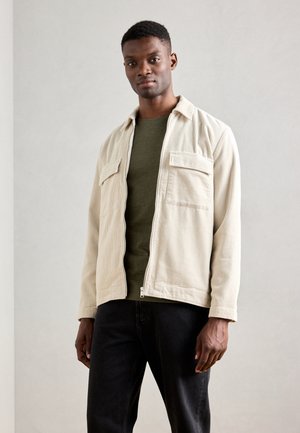 Cream corduroy jacket with two chest pockets, worn over a dark green t-shirt, paired with black jeans. Simple, functional design.