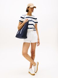 Camisa às riscas azul-marinho e branca com o logótipo "Tommy Hilfiger", calções brancos, boné bege e sapatilhas brancas com sola bege, a segurar uma bolsa de praia azul-marinho.