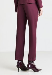 Donna che indossa pantaloni su misura di colore prugna e un giubbotto coordinato, abbinati a stivaletti a punta prugna lucidi con tacco a stiletto.