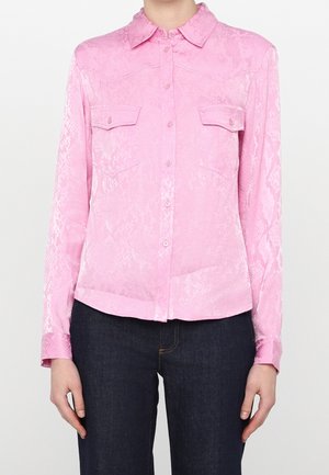 Kvinde iført en lyserød langærmet skjorte med knapper, med et diskret slangeskindsprint og forlommer med klapper, stylet med mørkeblå jeans.