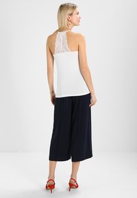 Valge tank top, millel on pitsdetail seljal, koos tumesiniste lühikeste pükstega ja punaste kontsadega sandaalidega. Sile kangas, läikiv disain ja ilma varrukateta.