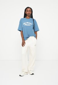 T-shirt oversized azul com "New Balance Athletics" em branco, combinado com calças de moletom creme com um logótipo discreto e ténis em tons neutros.