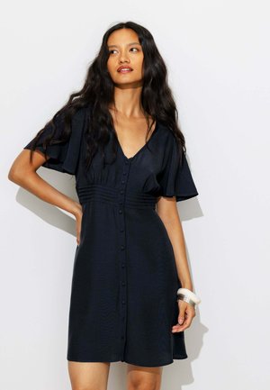 Femme aux longs cheveux ondulés portant une robe noire à manches courtes avec boutons sur le devant et des bracelets argentés, posant devant un fond blanc uni.