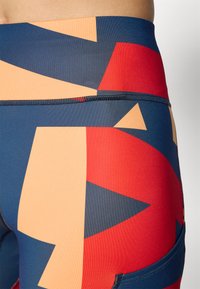 Barevné sportovní oblečení s geometrickým vzorem v červené, oranžové, námořnické a béžové barvě. Vyrobeno z pružného materiálu s elegantním designem a kapsou.