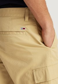 Shorts cargo beiges avec une poche latérale et un logo brodé comportant des accents rouges, blancs et bleus. Fabriqués à partir d'un matériau léger et lisse.