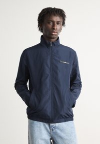 Veste zippée bleu marine avec un col haut, des poignets élastiques et une taille élastique. Présente un petit logo brodé sur la poitrine et deux poches latérales.