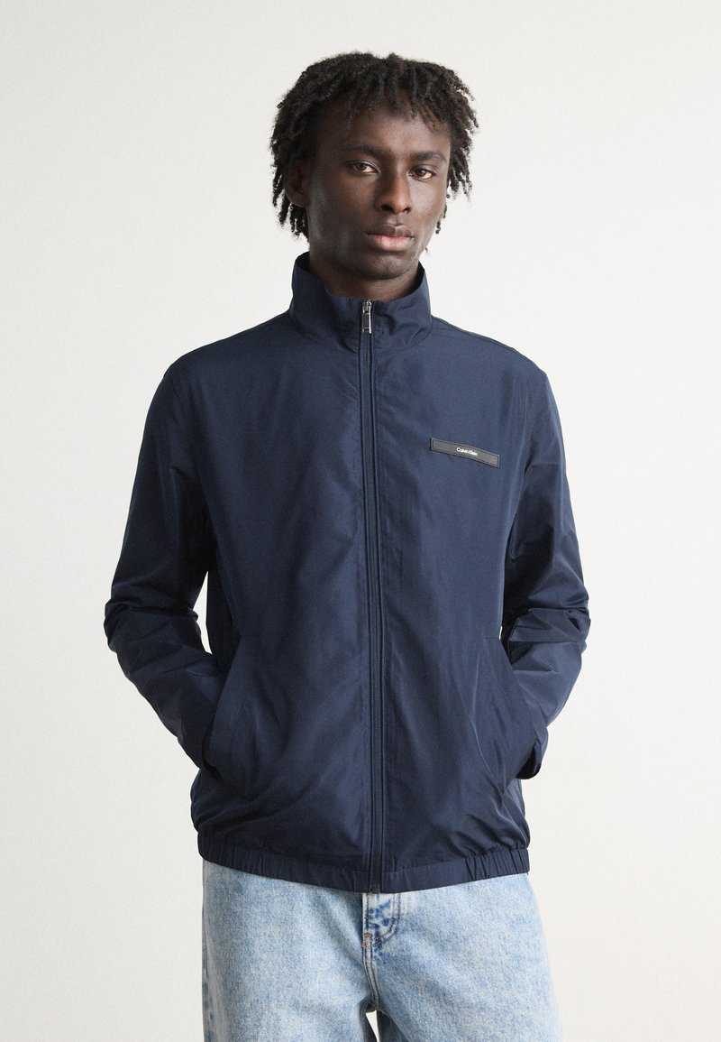 Veste zippée bleu marine avec un col haut, des poignets élastiques et une taille élastique. Présente un petit logo brodé sur la poitrine et deux poches latérales.