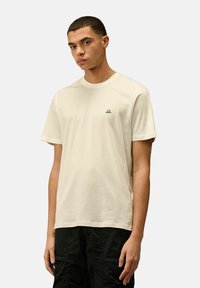 T-shirt di cotone color crema con scollo rotondo e maniche corte, con un piccolo logo nero sul lato sinistro del petto. Indossato con pantaloni neri.