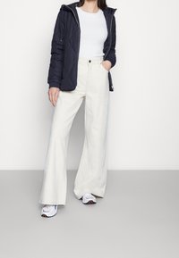 Granatowa pikowana kurtka z kapturem, biały żebrowany top i szerokie kremowe spodnie. Białe trampki z wielokolorowymi akcentami. Neutralne tło.