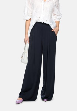 Woman wearing wide-leg black pants, white textured blouse with rolled sleeves, purple pointed shoes, and holding a white handbag.