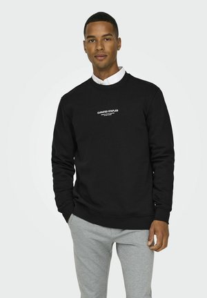 Zwarte sweatshirt met een ronde hals, lange mouwen, ribbelboorden en -zoom, met op de voorkant de witte tekst "CURATED STAPLES." Gecombineerd met lichtgrijze sweatpants.