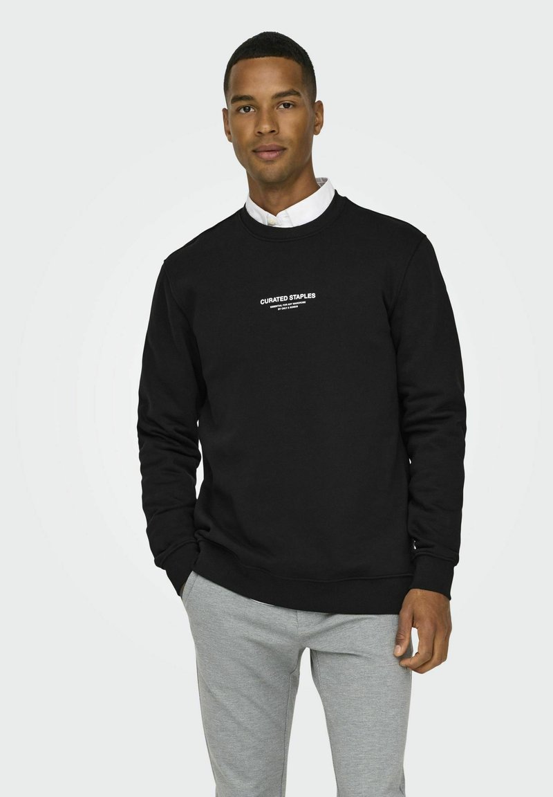 Schwarzer Sweatshirt mit Rundhalsausschnitt, langen Ärmeln, gerippten Bündchen und Saum, mit weißem Fronttext "CURATED STAPLES". Kombiniert mit hellgrauen Sweatpants.