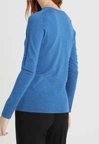 Blå långärmad tröja med rundad halsringning, med en mjuk textur och åtsittande design, bärs över svarta byxor.