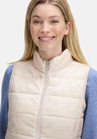 Beżowa pikowana kamizelka z wysokim kołnierzem, zapinana na zamek, z jasnoniebieską bluzką z długim rękawem pod spodem. Gładka faktura i minimalistyczny design.