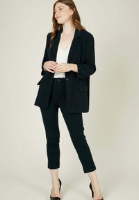 Blazer noir avec un revers cranté, pantalon fuselé assorti et un haut blanc à col en V. Le tissu semble lisse avec une coupe ajustée.