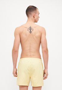 Κίτρινα μαγιό με ελαστική μέση, λεία υφή και πλαινές ραφές. Διαθέτει ένα μαύρο τατουάζ φλόρ-ντε-λίς στη μέση πλάτη.