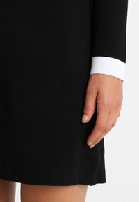 Ruka ženy odpočívá vedle černých dlouhých šatů s bílým manžetovým okrajem, ukazující část lemu šatů a horní část stehna.