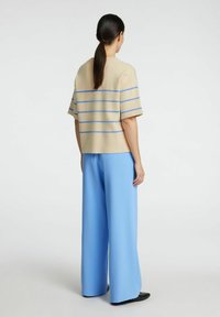 Top a maglia a righe beige con righe orizzontali blu, abbinato a pantaloni leggeri blu chiaro a vestibilità ampia e scarpe nere. Sfondo semplice.