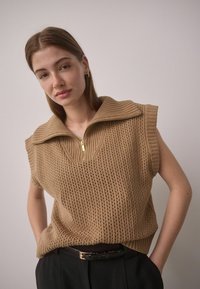 Tan sleeveless knit pullover with a high collar and zip closure. Features a textured open weave pattern and a relaxed fit. Black belt worn at the waist.