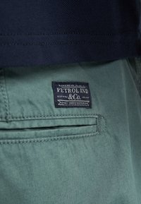 Gros plan sur la poche d'un pantalon vert avec une étiquette en tissu bleu foncé Petrol Ind & Co., partiellement recouverte par un vêtement bleu marine au-dessus.