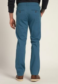 Tyrkysové chinos kalhoty s hladkou texturou, které mají dvě zadní kapsy s hnědými knoflíky a rovný střih, doplněné o hnědé boty.